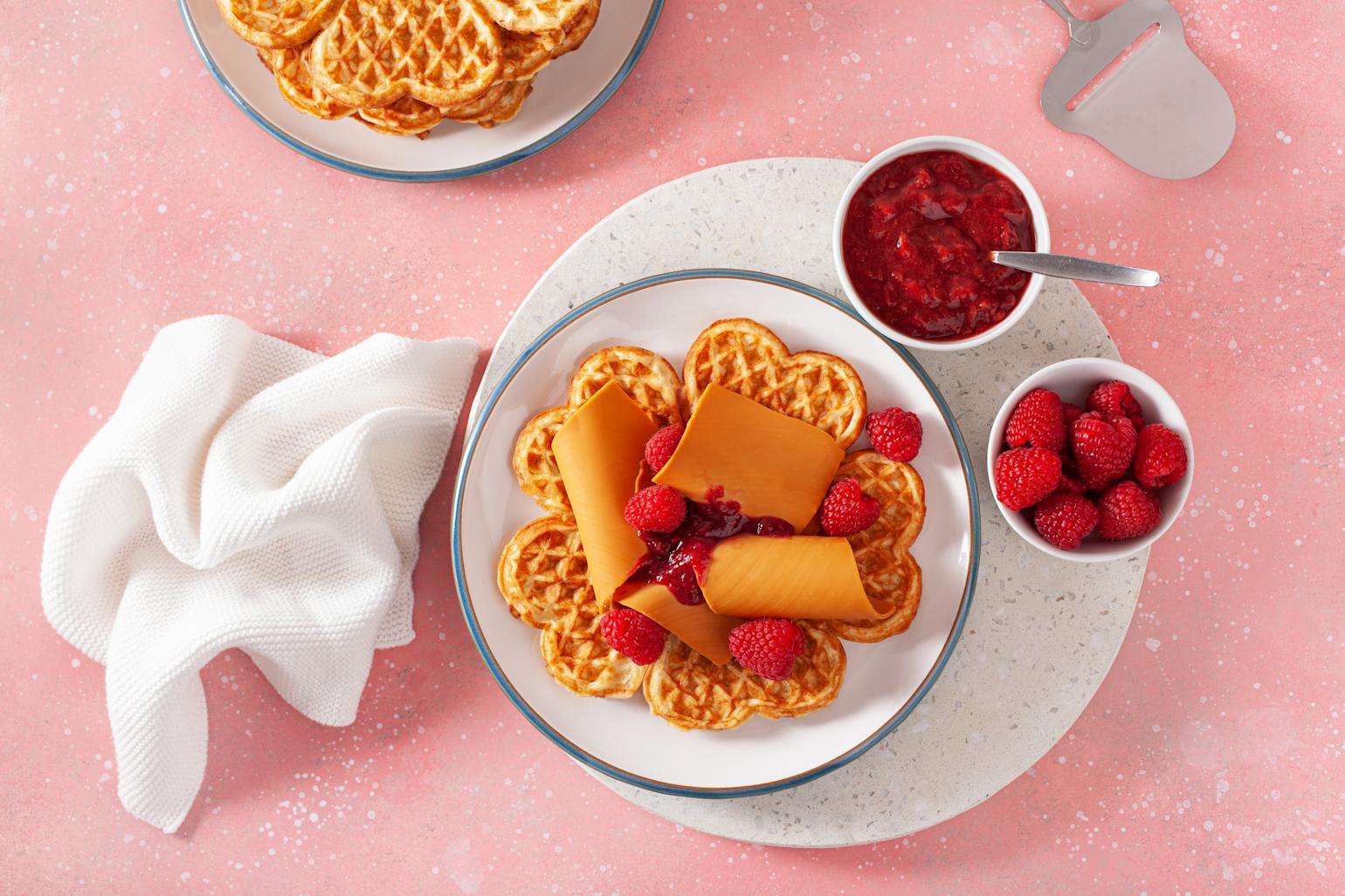 Vaffel på hvit tallerken, servert med brunostskiver, ferske bringebær og bringebærsyltetøy. Rosa bakgrunn.
