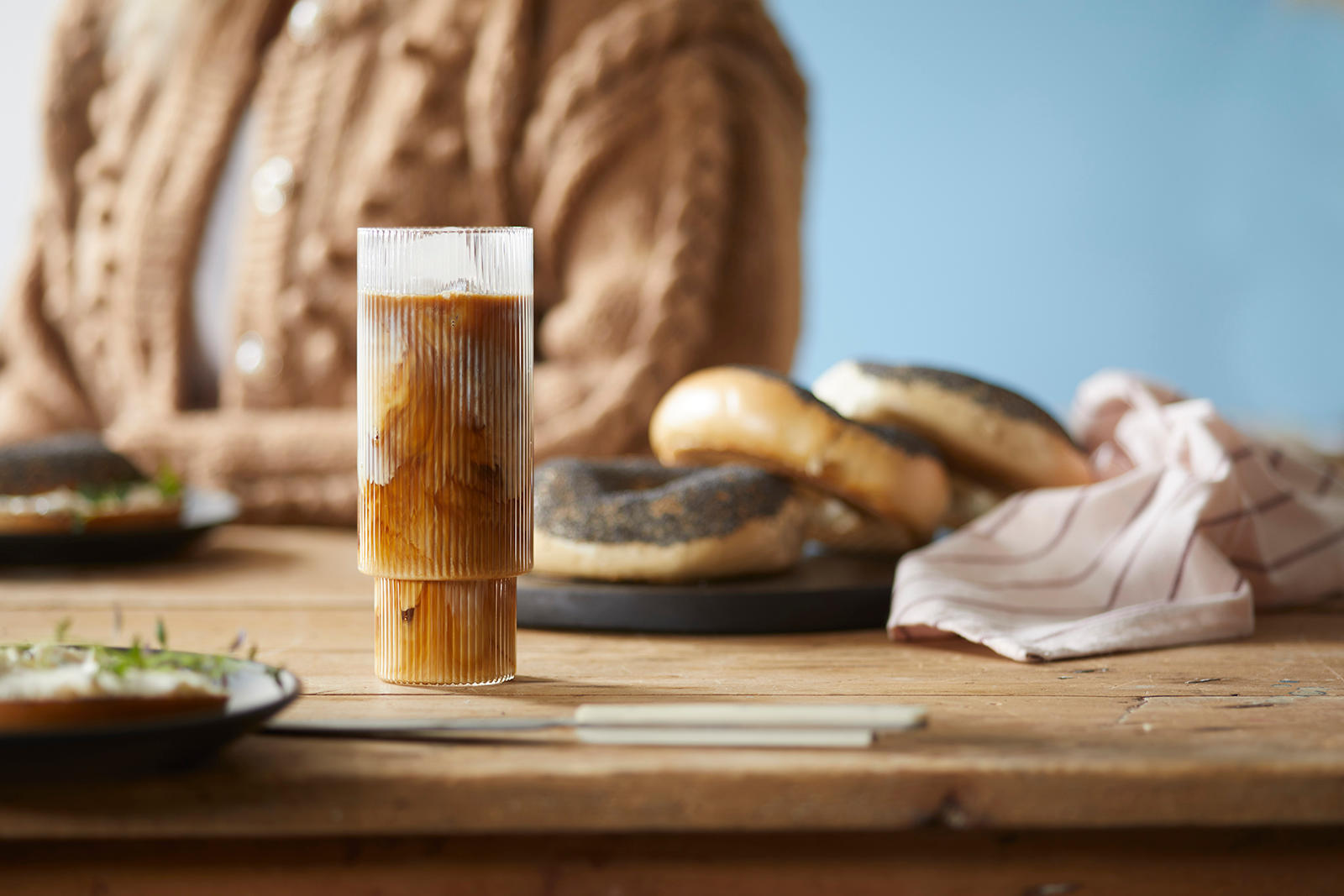 Iskaffe latte i høyt glass med isbiter, på dekket frokostbord. Brett med bagels og stripete kjøkkenhåndkle i bakgrunnen.