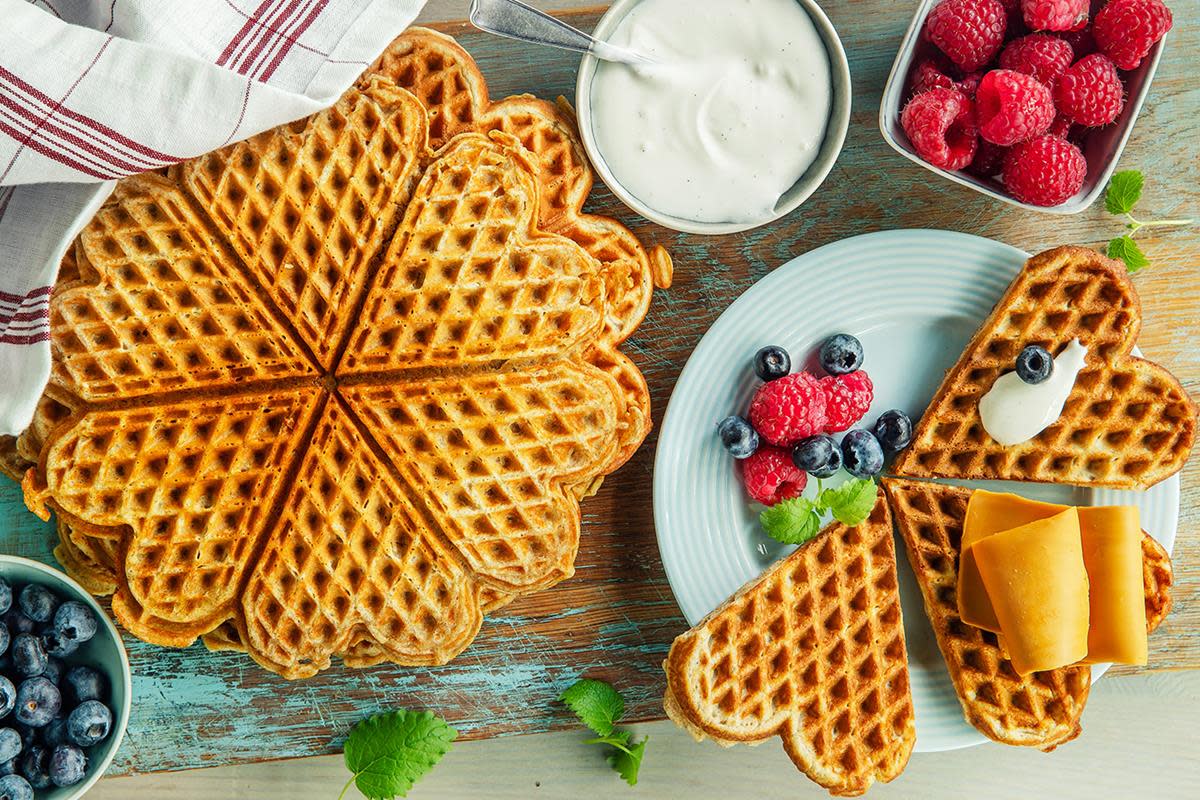 Stabel grove vafler under rødstripet kjøkkenhåndkle, står på rustikk trefjøl. På lyseblå asjett ved siden av er det vaffelhjerter med kesam, ferske bær og brunost. Ferske bær omkring.