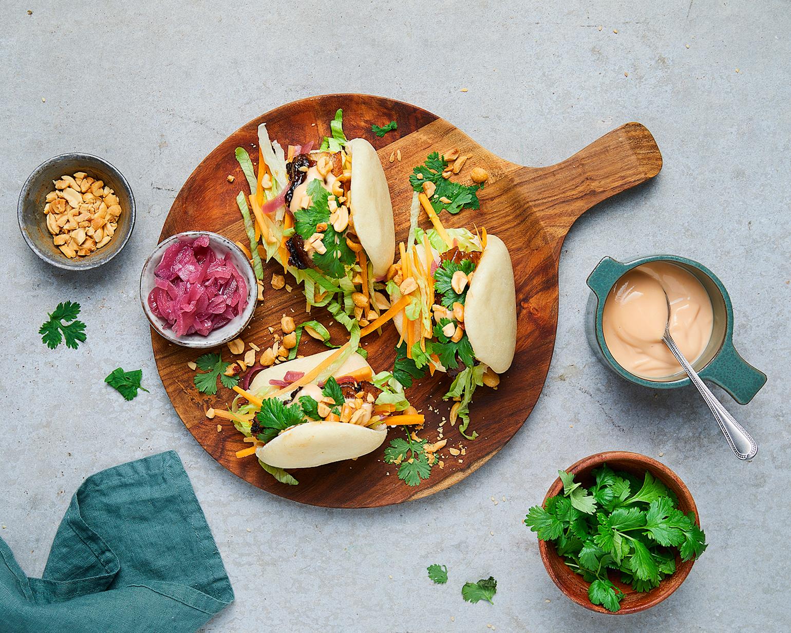 Tre steam buns med glasert ribbe og syltet rødløk servert på rund trefjøl, pyntet med frisk koriander og hakkede peanøtter. Små skåler med chilimayo, frisk koriander og hakkede peanøtter rundt fjøla.