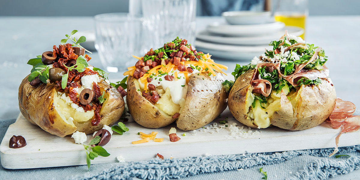 Bakt potet i tre varianter servert på hvit trefjøl. Til venstre med fetaost, soltørkede tomater og oliven. I midten med cheddar og sprøstekt bacon. Til høyre med parmesan, ruccola og fennikelsalami.