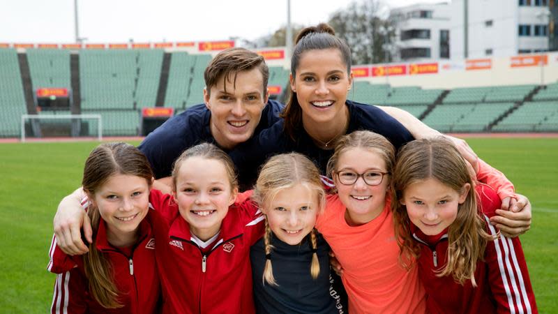 Karsten Warholm og Amalie Juul står bak fem barn og holder rundt dem på en friidrettsbane