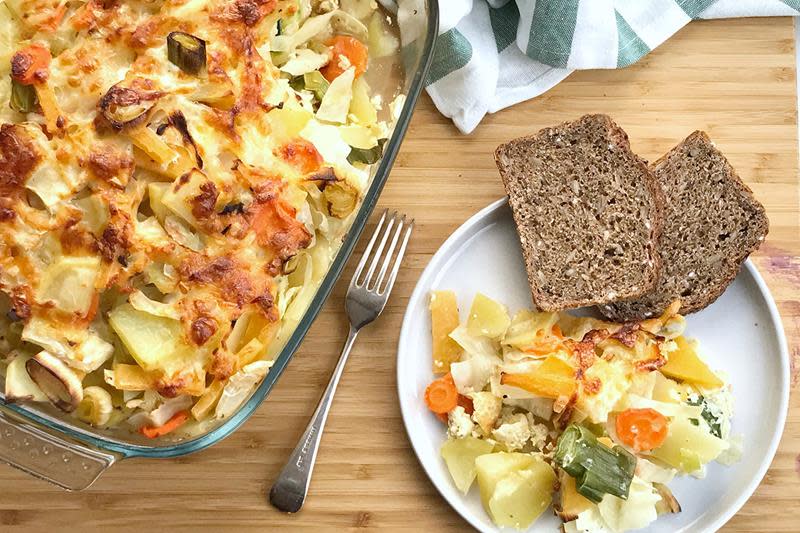 Sunn og saftig grønnsaksgrateng servert i ildfast form, og på hvit asjett ved siden av, med brød på siden. Alt står på lyst trebord.