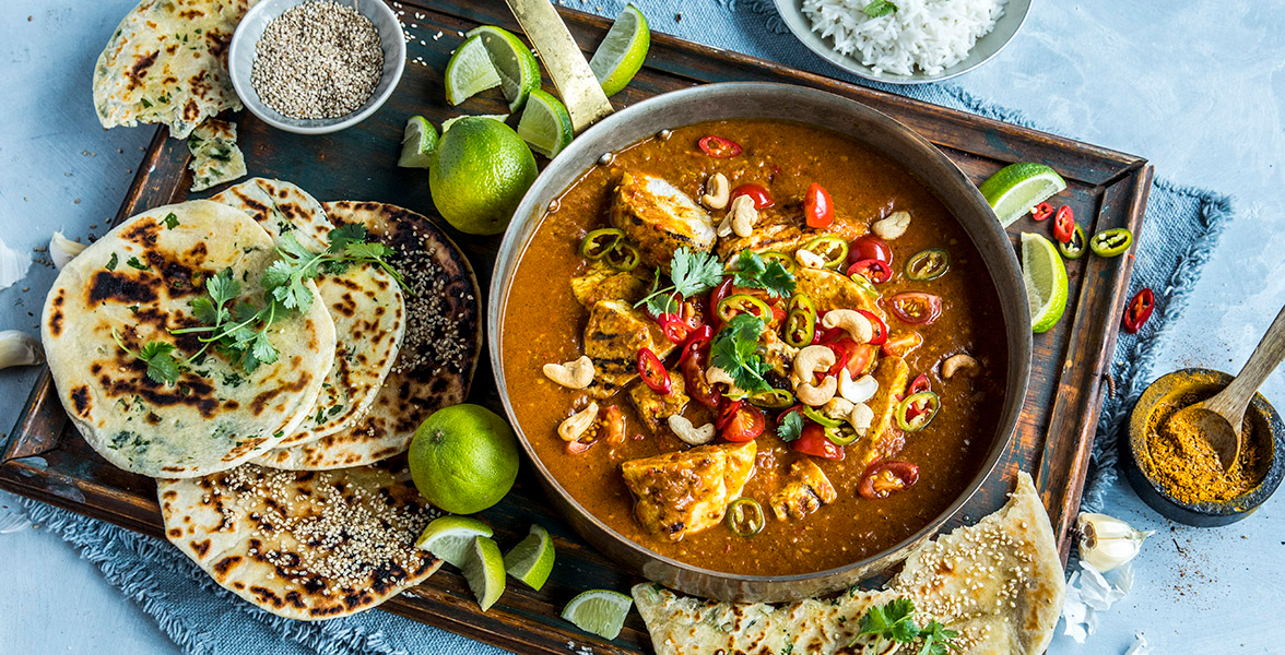 Kylling tikka masala servert toppet med cashewnøtter, frisk koriander og frisk chili i en metallgryte på trefjøl. Limebåter og hel lime, og naanbrød rundt.