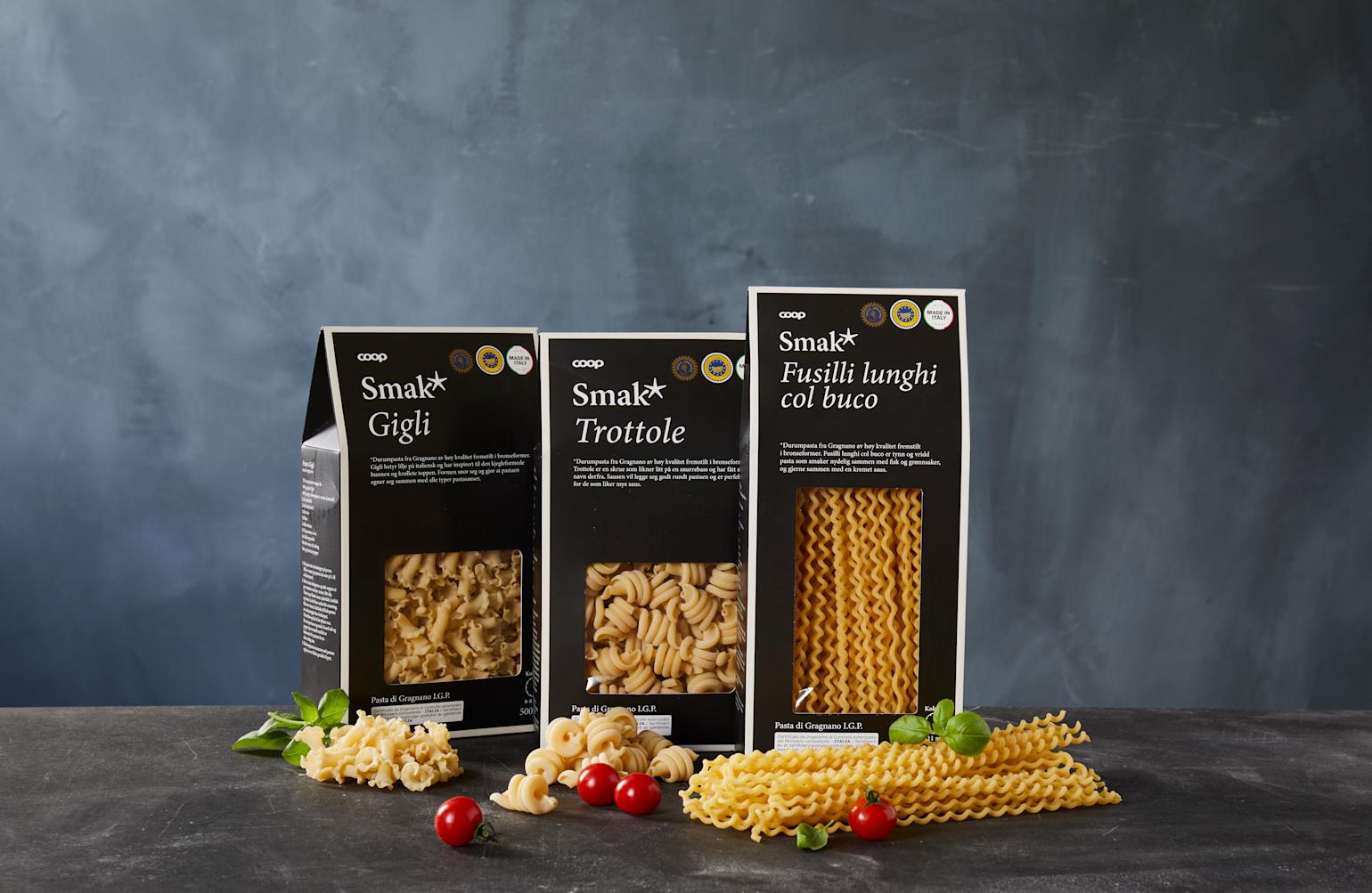 3 forskjellige pakker med Smak* pasta, med de forskjellige typene pasta lagt ut på mørkt bord foran pakkene.