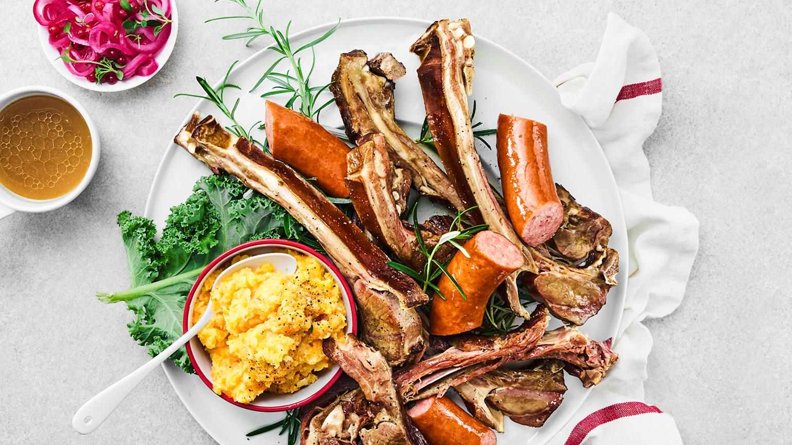 Pinnekjøtt i grønnsakskraft med klassisk tilbehør servert på hvit tallerken med rotmos, vossakorv og frisk rosmarin. Små skåler med grønnsakskraft og syltet rødløk ved siden av.