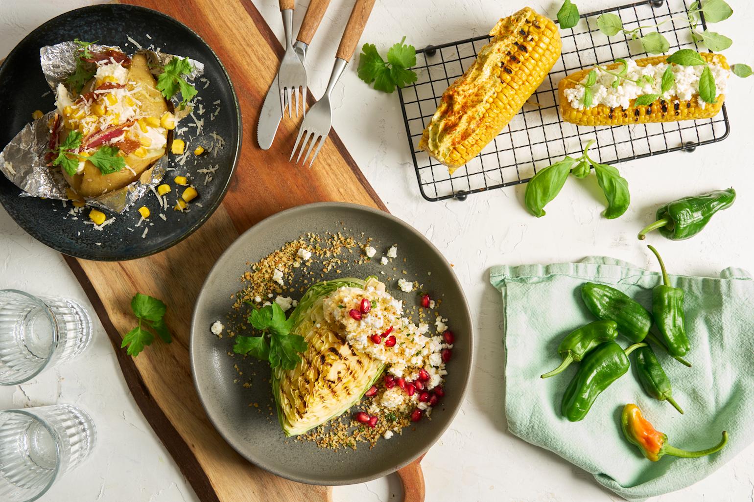 Coop Grill Perfekt bakepotet, nykål, maiskolber og padron servert på asjetter, rist og turkis linserviett. Maiskolbene har kryddersmør og fetakrem på toppen, bakt potet er fylt med rømme, mais og bacon, og nykålen har fetakrem.