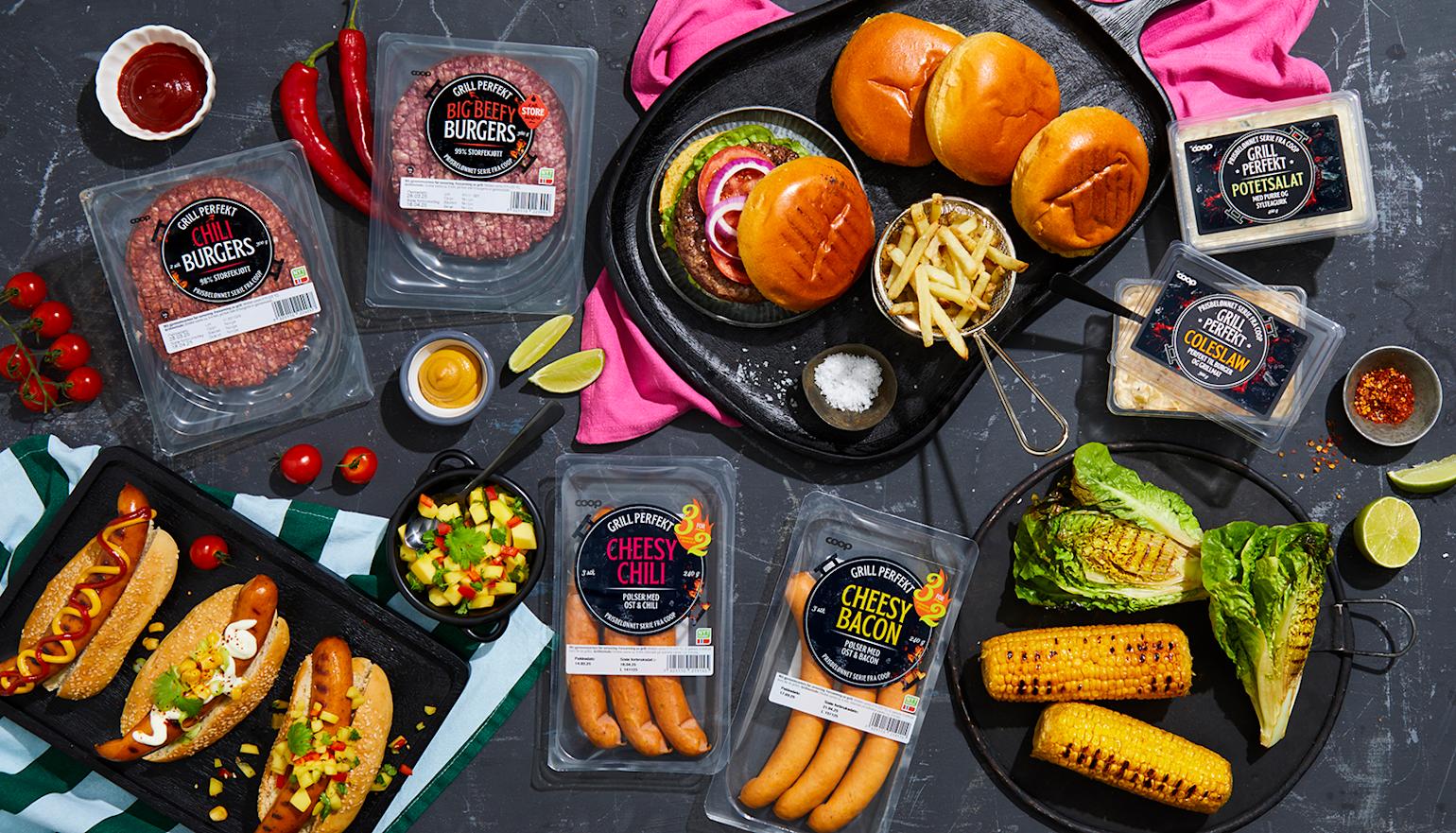 Grill Perfekt burgere og pølser i emballasje på mørk steinbakgrunn, samt tilberedt på svarte serveringsfat. Grillede maiskolber og grillet hjertesalat, og Grill Perfekt tilbehør ved siden av.