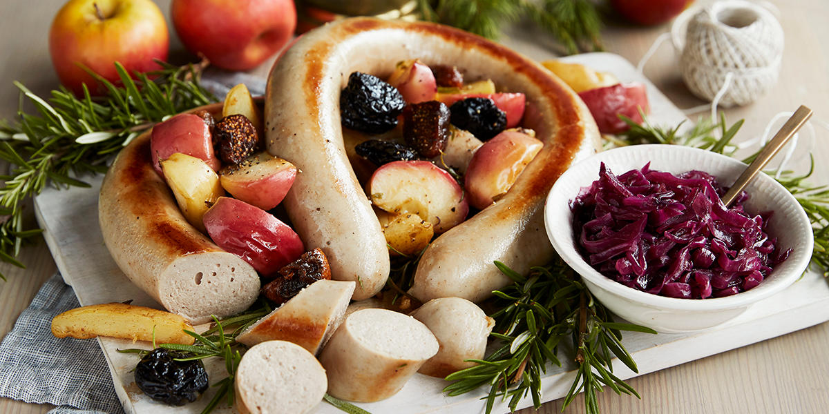 Julepølse med stekte epler og rødkålkompott servert på lys trefjøl med fiken, friske urter, og granbar til pynt. Liten hvit skål med rødkålkompott på siden.