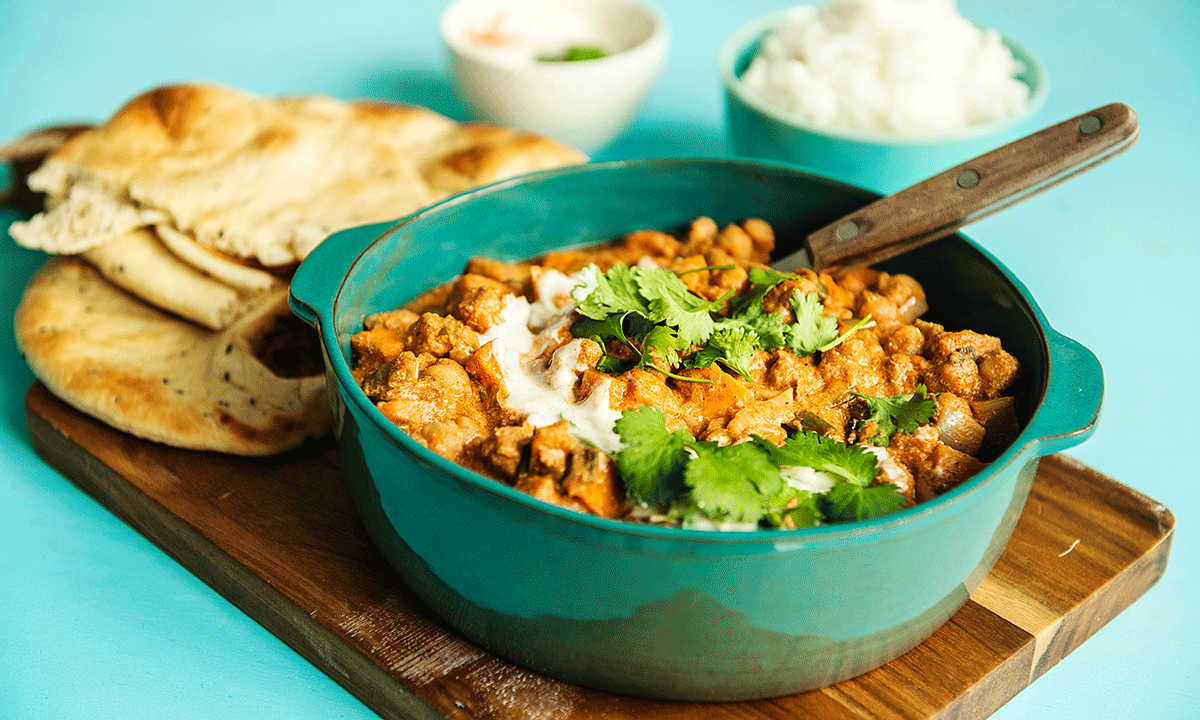 Vegetar tikka masala med søtpoteter og kikerter i høy grønn tallerken. Ris og nanbrød i bakgrunnen.