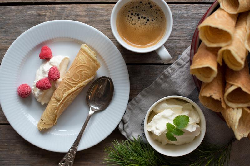 Hjemmelagde sprø krumkaker i kjegleform servert på hvit asjett med pisket krem, ferske bringebær og en kopp kaffe. Asjetten står på rustikk trebord, med et fat krumkaker og en liten skål pisket krem ved siden av.