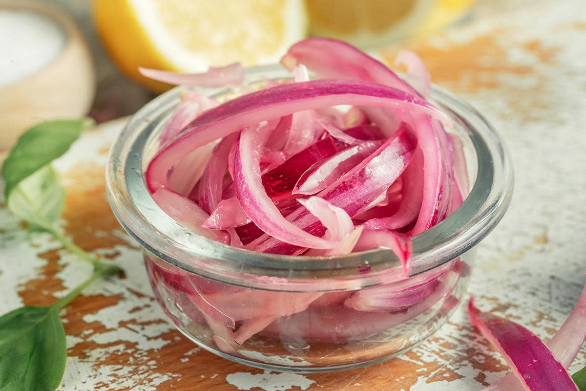 Glass med knall rosa syltet rødløk i strimler, servert på rustikk trefjøl. Salt, ferske urter og delt sitron i bakgrunnen.