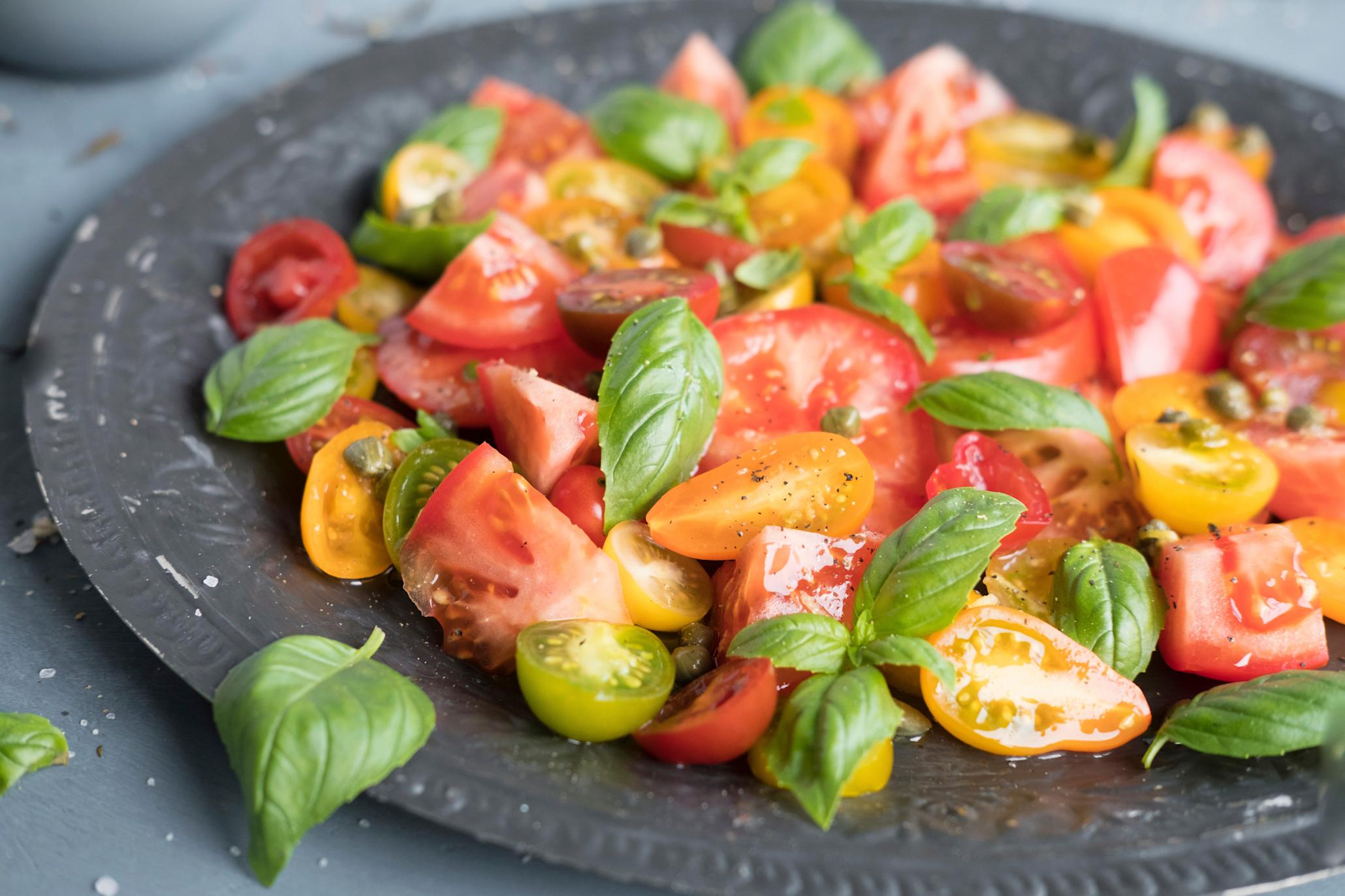 Fargerik tomatsalat med kapers, og tomater i gult, rødt og grønt, servert på mørkegrå tallerken. Tomatsalaten er toppet med fersk basilikum og syrlig oljedressing.