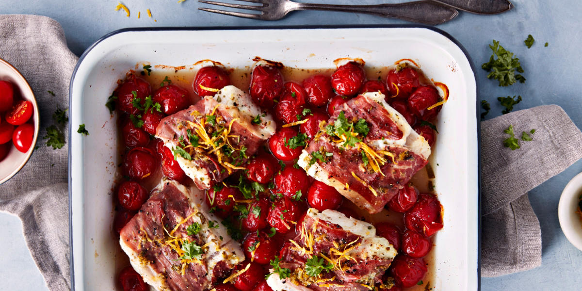 Ovnsbakt skrei med spekeskinke og cherrytomater servert i hvit ildfast form med ferske urter og sitronskall revet over. Formen står på et blått bord med linduk og bestikk.