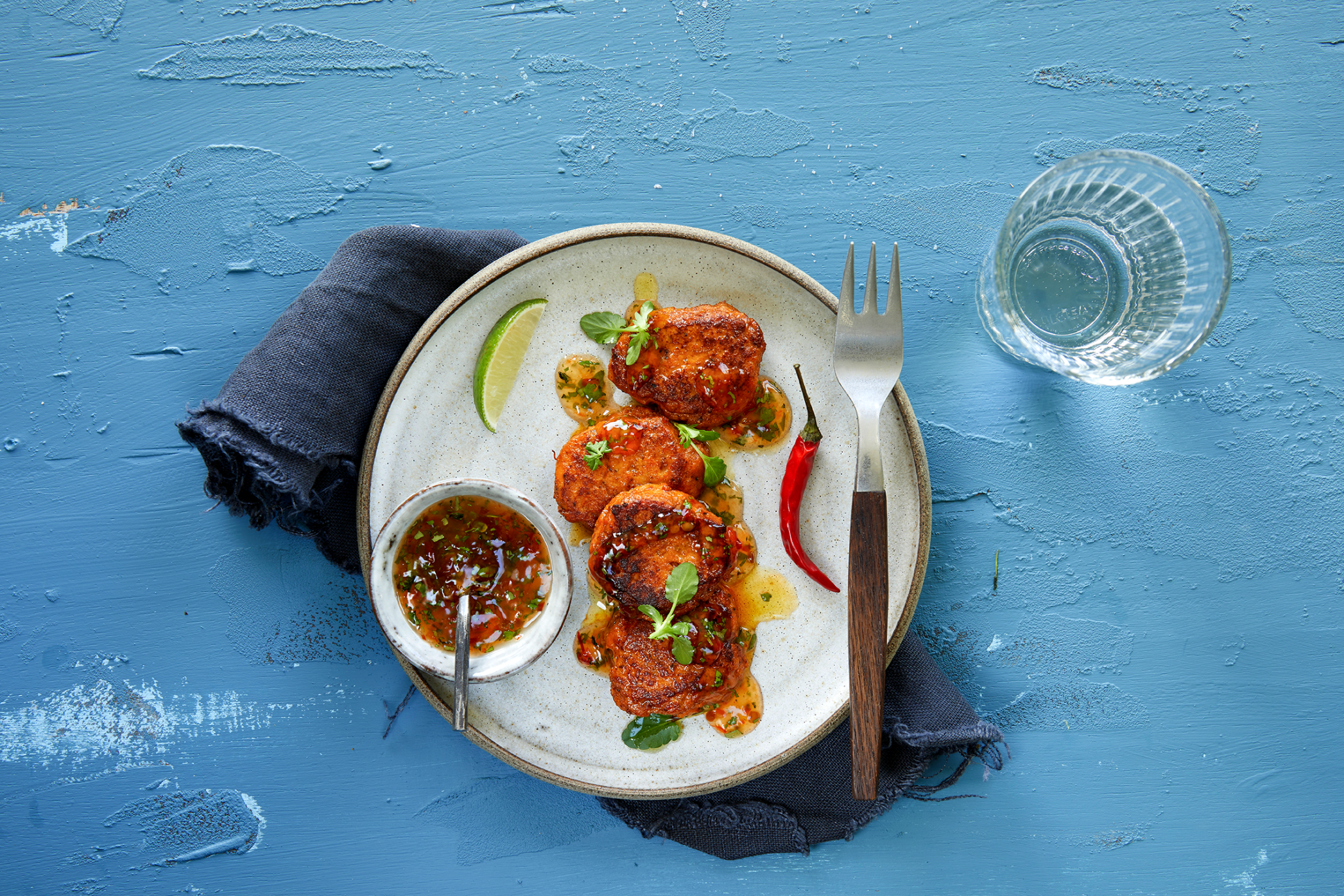 Fiskekaker med inspirasjon fra Thailand. Fiskekaker laget av laks servert på lys tallerken med chili, lime, ferske urter og dip i en skål.