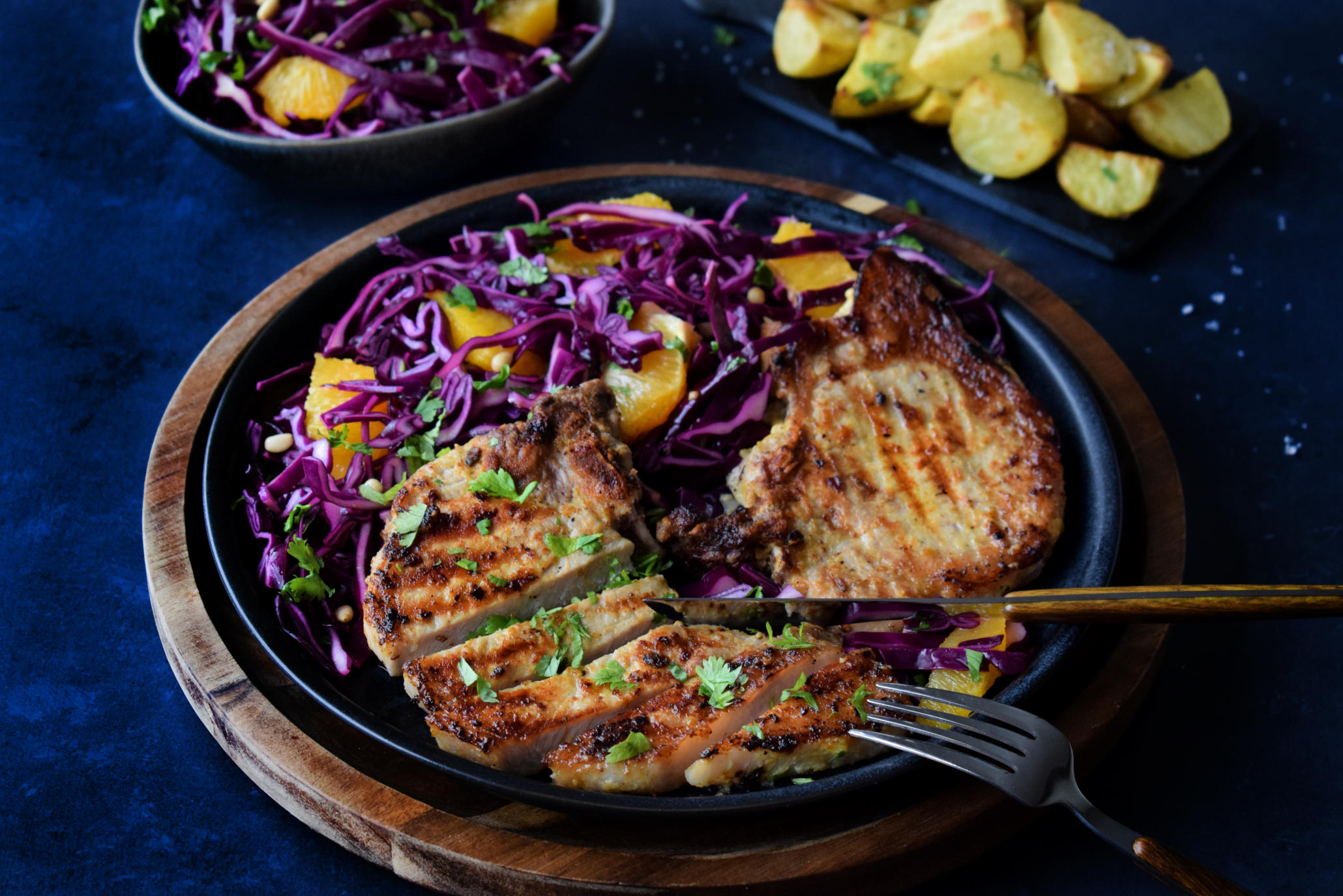 Sennepmarinerte sommerkoteletter med frisk rødkålslaw med appelsin på mørk tallerken, med mørkt trefat under. Grillpoteter og skål med rødkålslaw i bakgrunnen.