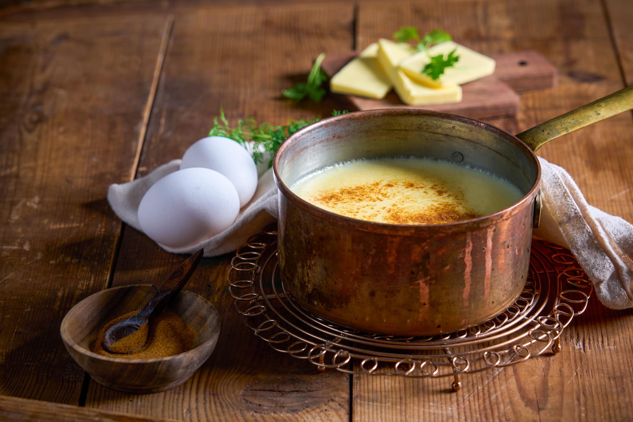 Hollandaise saus servert i liten messingkjele på messingrist, på rustikk trebord. Kjelen er omringet av nøkkelingrediensene i Hollandaise; egg og smør. Liten treskål med kajennepepper på siden.