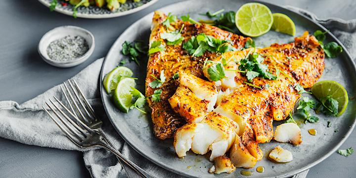 Spicy ovnsbakt skrei med krydret marinade servert på grå tallerken med koriander og limebåter. Tallerken med mango- og avokadosalat i bakgrunnen.