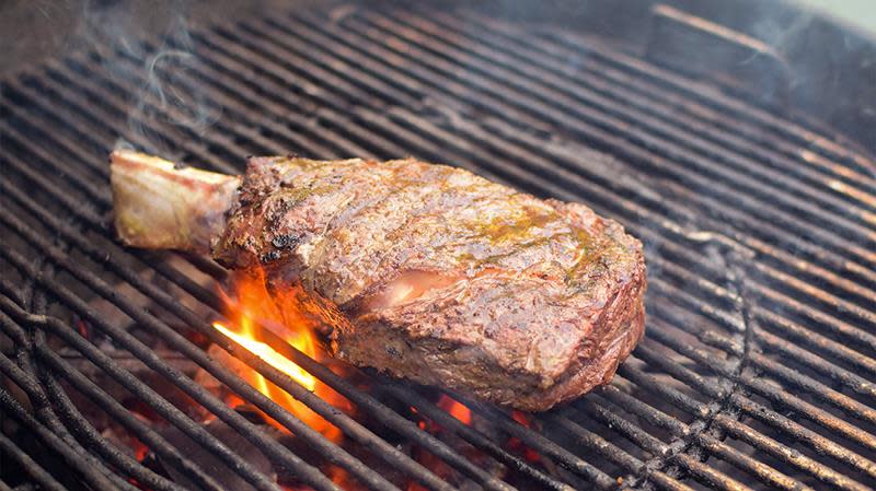 Grillet tomahawk steak på kullgrill, med flammer og røyk rundt kjøttet.