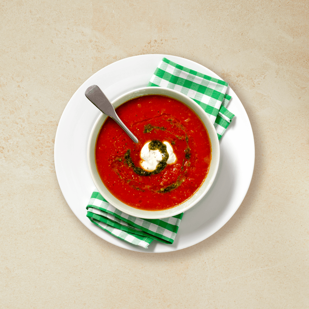 Tomatsuppe med pesto og rømme, servert i en hvit skål på et grønt og hvitt rutete håndkle som ligger på en hvit tallerken