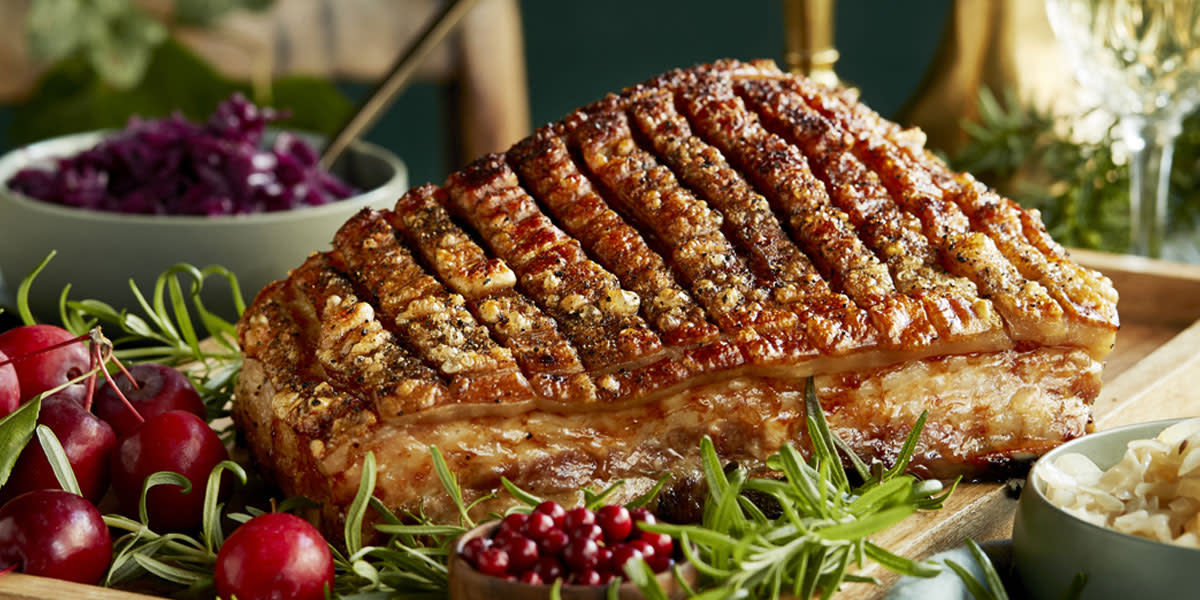 Langtidsstekt ribbe med sprø svor, servert på serveringsbrett i tre på dekket julebord, med fersk rosmarin, liten skål med tyttebær og røde plommer ved siden av. På siden står to skåler med rødkål og surkål.