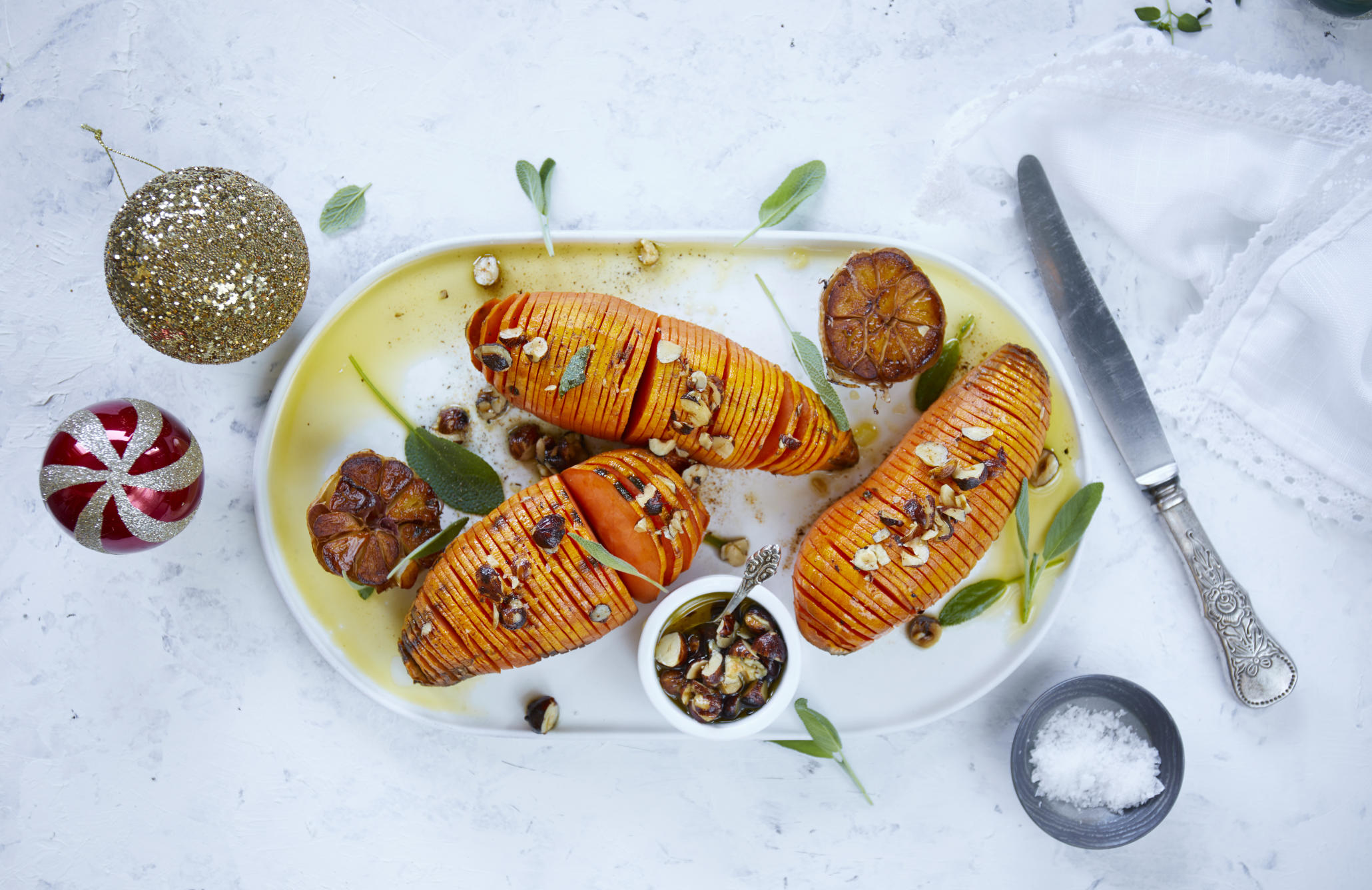 Hasselback søtpoteter på fat, med stekt hel hvitløk delt i to, overstrødd med hasselnøtter og ferske urter.