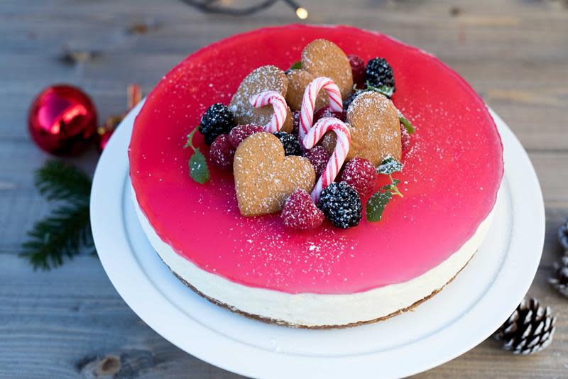 Julepyntet ostekake med rødt gelelokk, toppet med hjertepepperkaker, bringebær, bjørnebær, candy canes, sitronmelisse og et dryss melis. Står på rustikk trebakgrunn, omringet av julelys, kongler, granbar og en rød julekule.