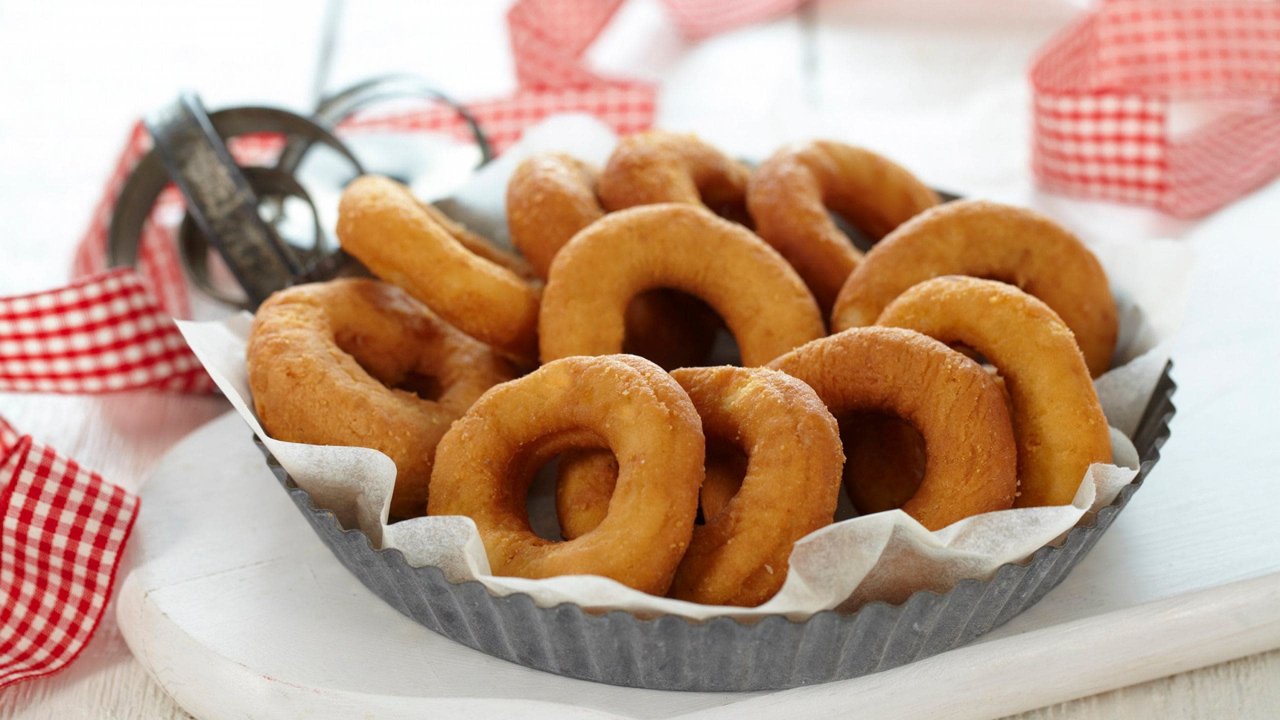 Glutenfrie smultringer servert i rund metallform med hvitt bakepapir. Smultringformer og rød -og hvitrutete bånd i bakgrunnen.